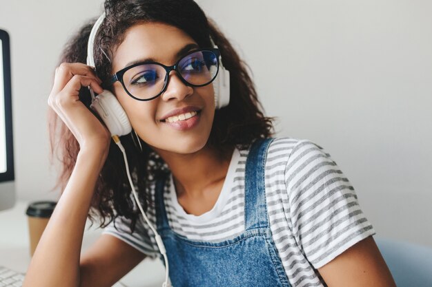 Inspiriertes brünettes Mädchen mit gebräunter Haut, die lacht, während Musik im Büro hört