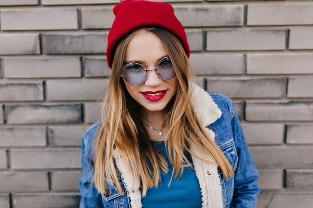 Inspiriertes kaukasisches Mädchen mit roten Lippen, die draußen mit freudigem Lächeln aufwerfen. Raffiniertes weibliches Modell in Jeansjacke, die während des Frühlingsspaziergangs entspannt.