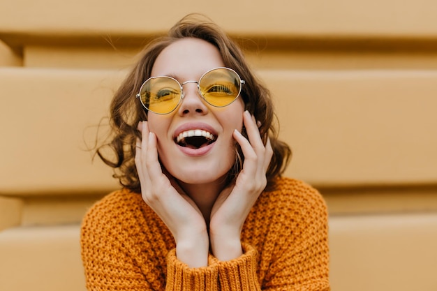 Inspiriertes kurzhaariges Mädchen in gelber Brille, das mit überraschtem Lächeln vor dem Gebäude posiert. Nahaufnahmefoto im Freien von erstaunlicher stilvoller Frau in Strickpullover, die lacht und wegschaut.