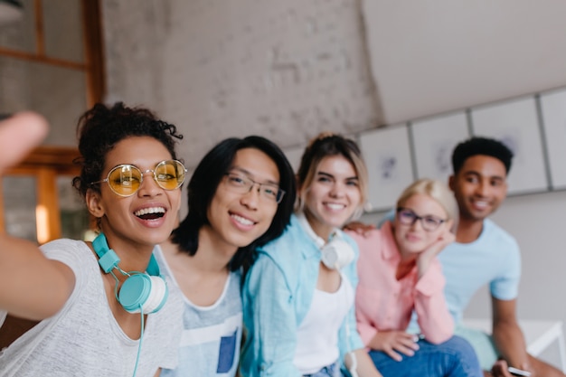 Kostenloses Foto inspiriertes mädchen in stilvollen gelben gläsern, die selfie mit ihrem asiatischen universitätsfreund und anderen studenten machen. charmante junge frau mit hellbrauner haut, die foto von sich mit leuten macht.