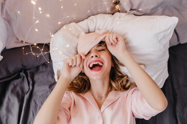 Inspiriertes Mädchen mit lockiger Frisur, die im Bett lacht. Innenaufnahme der faszinierenden Dame, die mit geschlossenen Augen auf weißem Kissen aufwirft.