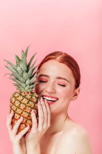 Inspiriertes weibliches Modell, das Ananas hält. Studioaufnahme des lachenden Ingwermädchens mit der Frucht, die auf rosa Hintergrund aufwirft.