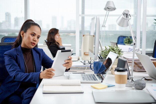 Intelligent gekleidete Asiatin, die am Schreibtisch im Büro mit Tablette sitzt