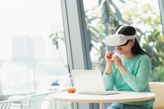 Intelligente, attraktive asiatische Geschäftsinhaberin, die eine Virtual-Reality-Brille trägt, genießt ein zwangloses Metaverse-Treffen mit Konzentrat im Café-Restaurant