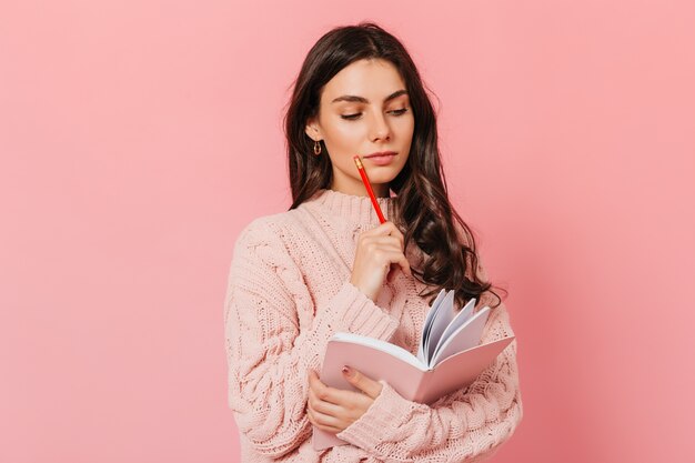 Intelligente Frau im rosa Pullover denkt über die Fortsetzung ihres Buches nach. Brünette schaut in ihrem Tagebuch auf rosa Hintergrund.