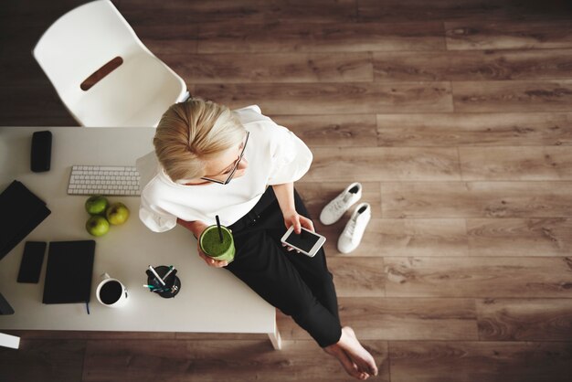 Intelligente Geschäftsfrau mit Smoothie und Smartphone am Schreibtisch sitzend