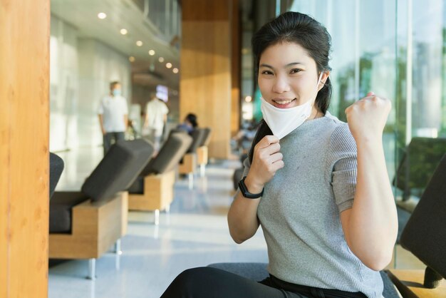 Intelligente gesunde asiatische Frauen tragen Gesicht Covid 19 Schutz sitzen entspannen Hand aufstehen besser starker Kampf Virus verbreitet soziale Distanzierung im Hintergrund des Krankenhauses im öffentlichen Wartebereich