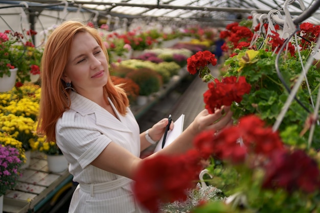 Intelligente Gewächshaussteuerung. Arbeitnehmerin inspiziert rote Blumen und notiert Daten bei Tageslicht
