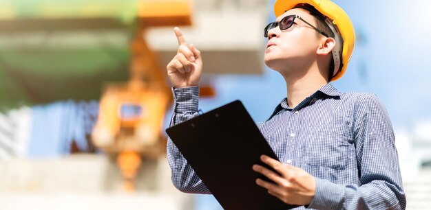Intelligenter asiatischer männlicher professioneller Bauingenieur hält Klemmbrett-Baustellenprüfung mit Infrastruktur- und Gebäudehintergrund