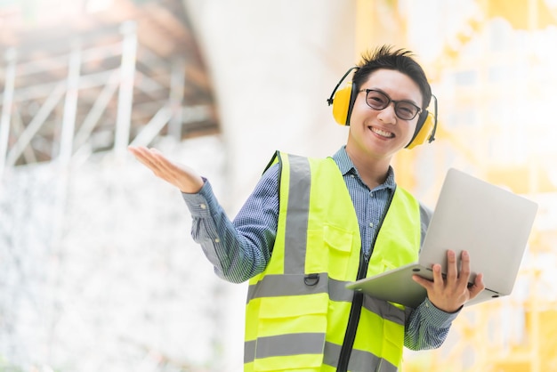 Intelligenter asiatischer männlicher professioneller Bauingenieur hält Laptop-Standort-Baukontrolle mit Infrastruktur- und Gebäudehintergrund