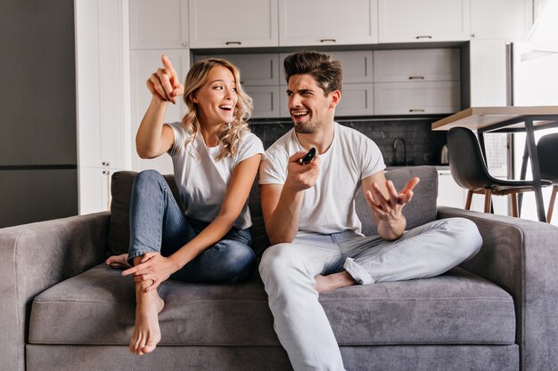 Interessierte blonde Dame fernsehen. Innenporträt des lächelnden Paares, das auf gemütlicher Couch sitzt.