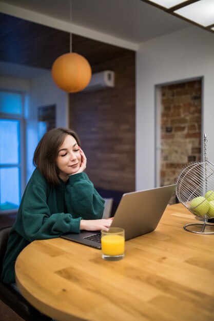 Interessierte brünette Frau arbeitet an ihrem Laptop auf dem Küchentisch, der Orangensaft trinkt