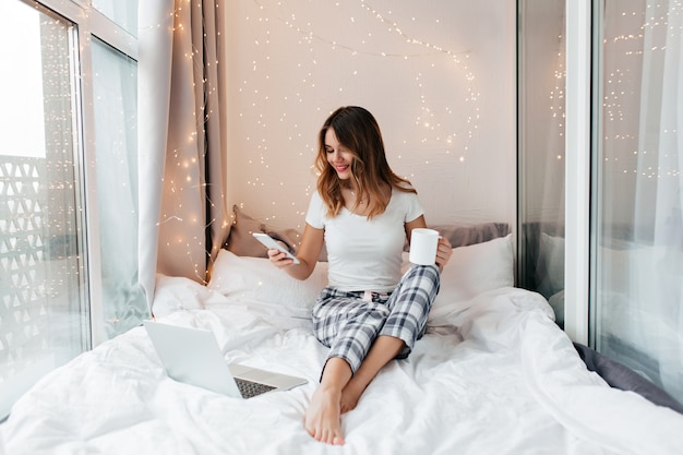 Interessierte europäische Frau, die am Morgen mit Laptop und Tasse heißem Kaffee kühlt. Innenfoto des bezaubernden Mädchens, das im Bett sitzt und SMS sendet.