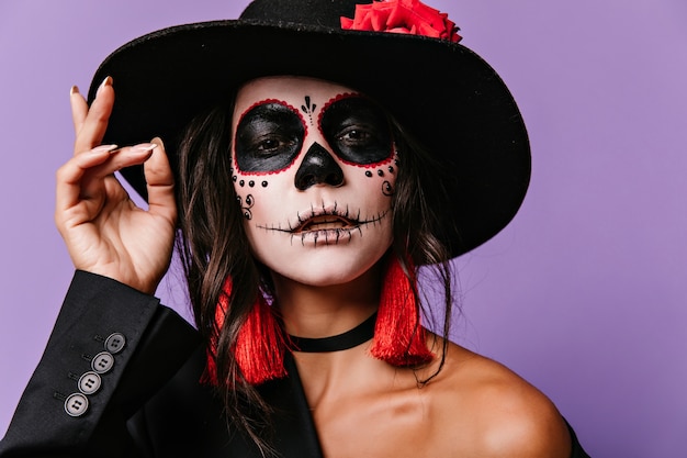 Interessierte Frau mit gruseliger Gesichtsbemalung. Halloween-Porträt des brünetten lateinamerikanischen Mädchens im großen schwarzen Hut.