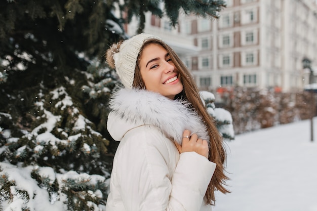 Interessierte langhaarige Frau in weißer Kleidung, die glückliche Winterzeit genießt und lacht. Außenporträt der prächtigen europäischen Frau in der Strickmütze, die an der verschneiten Straße steht
