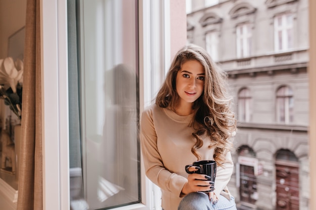 Interessierte schüchterne Frau mit langen Haaren, die mit Tasse Tee auf Schwelle aufwirft