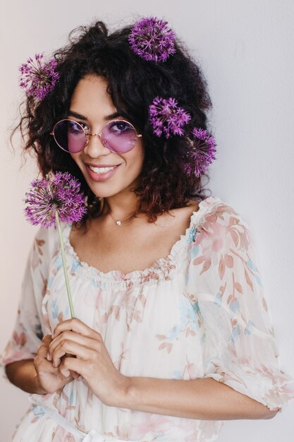 Interessierte schwarze Frau mit stilvollem Haarschnitt, der mit lila Blume aufwirft. Atemberaubendes afrikanisches weibliches Modell, das Allium hält und schaut.