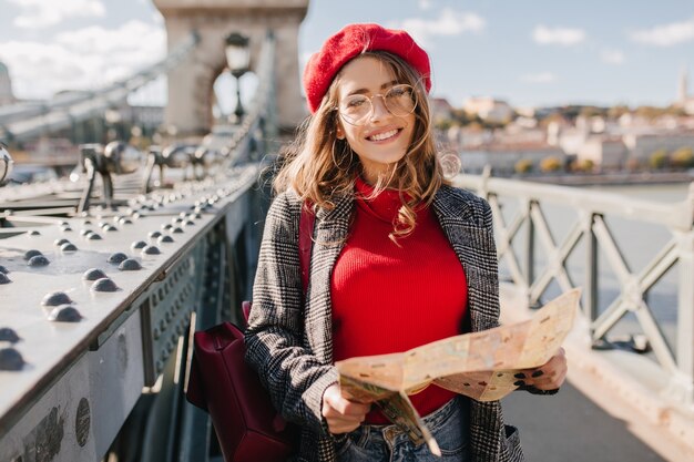 Interessierte weiße Frau im roten Pullover und in der Baskenmütze, die Zeit im Freien verbringen und Stadt mit Karte erkunden