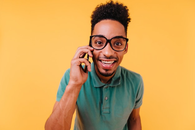 Interessierter dunkeläugiger Mann in Brille, der am Telefon spricht. Glücklicher afrikanischer Kerl trägt grüne Kleidung, die mit Smartphone aufwirft.