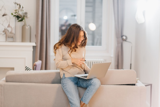 Interessiertes lockiges Mädchen mit Laptop, auf Sofa sitzend und lachend