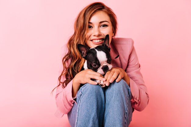 Interessiertes Mädchen mit gewellter Frisur, das Freizeit mit Hund verbringt. Porträt der jungen Frau, die mit französischer Bulldogge aufwirft.