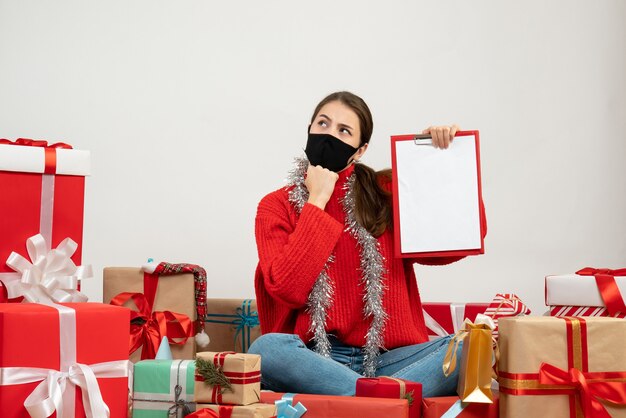 Interessiertes Mädchen mit schwarzer Maske, die Dokumente hält, die um Geschenke auf Weiß sitzen