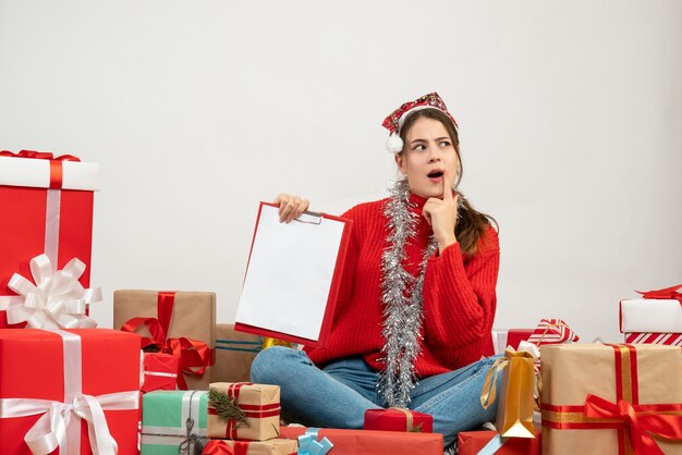 Interessiertes Mädchen mit Weihnachtsmütze, das Dokument hält, das um Geschenke auf Weiß sitzt