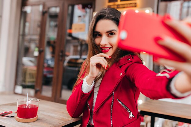 Interessiertes süßes Mädchen, das Tee trinkt und ein Foto von sich macht. Outdoor-Porträt der sinnlichen jungen Frau in roter Jacke mit Telefon für Selfie.