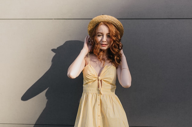 Interessiertes weibliches Ingwermodell, das ihren Strohsommerhut während des Fotoshootings berührt. Foto der attraktiven rothaarigen Frau im gelben Kleid, das sich in gutem Tag entspannt.