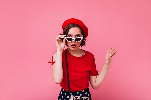 Interessiertes weißes Mädchen mit kurzen Haaren, die durch Brille schauen. prächtige Französin im roten Outfit.