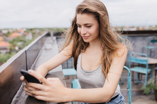 Interessiertes weißes Mädchen mit Smartphone auf der Straße. Außenaufnahme der sorglosen langhaarigen Frau, die Nachricht während des Kühlens im Dachcafé tippt.