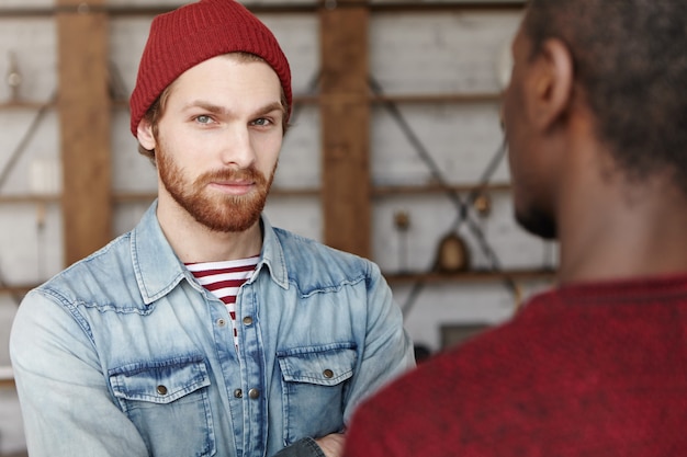 Kostenloses Foto interracial freundschafts- und partnerschaftskonzept. zwei beste freunde treffen sich im café und besprechen pläne und ideen ihres gemeinsamen vielversprechenden geschäftsprojekts. weißer bärtiger mann mit hut