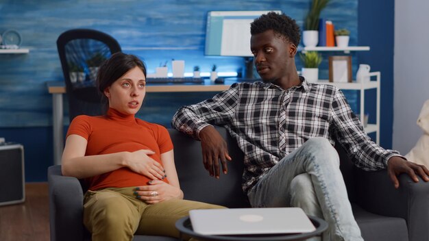 Interracial Paar mit Schwangerschaft entspannt auf der Couch zu Hause. Multiethnische Eltern sprechen über Kind und Elternschaft, während ein afroamerikanischer Mann den Babybauch einer kaukasischen Frau berührt