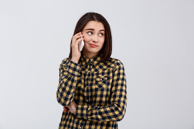 Ironisches junges schönes Mädchen gekleidet im karierten Hemd, das am Telefon spricht und Seite über weißer Wand betrachtet.