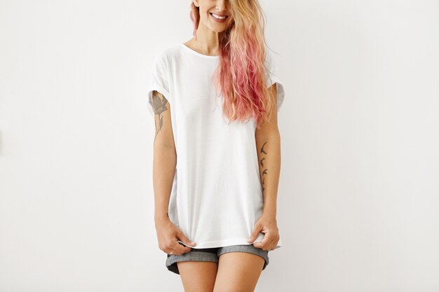 Isolierte abgeschnittene Aufnahme der lächelnden fröhlichen jungen Frau mit Tätowierungen auf Armen und unordentlicher rosa Frisur stehend, gekleidet in Jeansshorts und weißem T-Shirt