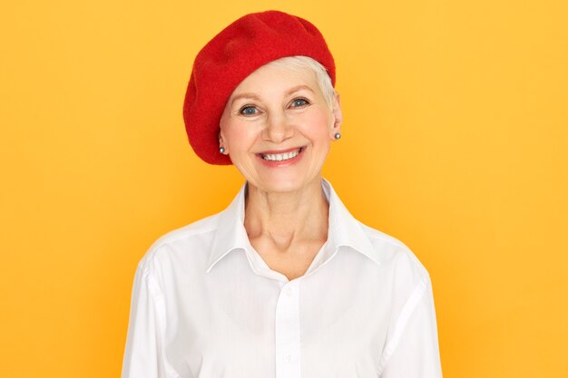 Isolierte Aufnahme der glücklichen gut aussehenden positiven Frau mittleren Alters mit kurzen Haaren, die gegen gelben Hintergrund aufwirft, rote Haube und weißes Hemd tragend, Kamera mit fröhlichem breitem Lächeln betrachtend