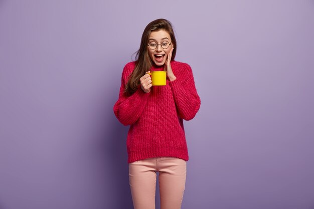 Isolierte Aufnahme der glücklichen überglücklichen Frau schaut auf Tasse des heißen aromatischen Getränks, hält gelben Becher, trägt Brille, roten Pullover, steht gegen lila Wand. Freudige Frau hat Kaffeepause. Trinkkonzept