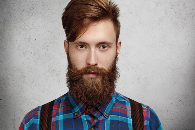 Isolierte Aufnahme des attraktiven modischen kaukasischen Mannes mit dem unscharfen Hipster-Bart und dem Schnurrbart, die das karierte Hemd tragen, das mit ernstem und nachdenklichem Ausdruck schaut, während an etwas gedacht wird