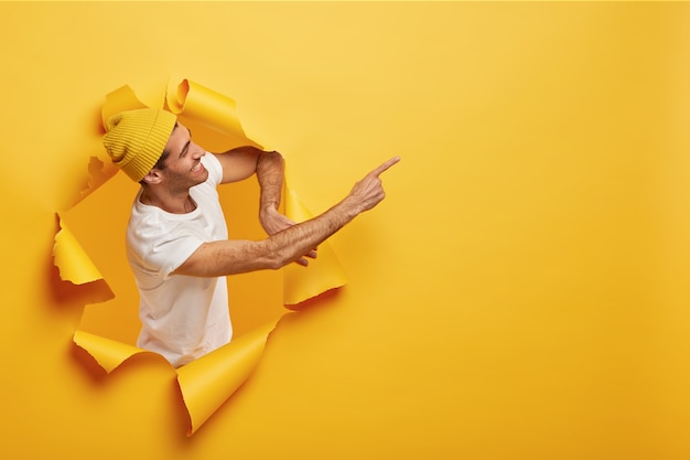 Isolierte Aufnahme des zufriedenen männlichen Modells steht seitlich im Papierloch, gekleidet in gelbe Kopfbedeckung