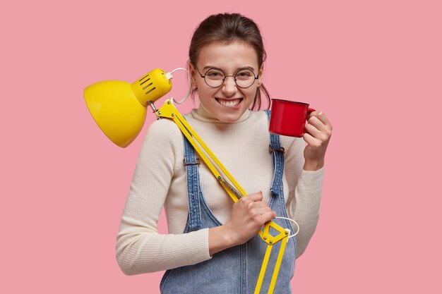 Isolierte Aufnahme von attraktiven lächelnden Frau beißt Unterlippe, trägt runde Brille, Jeansoverall, hat Spaß während der Kaffeepause, trägt Lampe