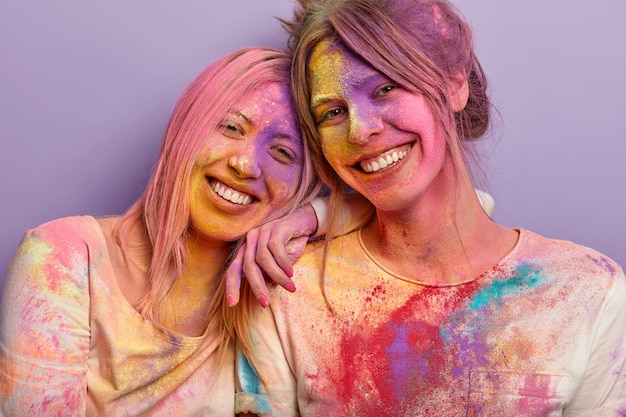 Isolierte Aufnahme von freundlichen optimistischen Mädchen, die müde sind, nachdem sie farbiges Pulver auf die Gesichter geschmiert haben, stehen eng, feiern kommenden Frühling während Holi-Feiertag. Aufnahme von zwei Frauen in leuchtenden Farben