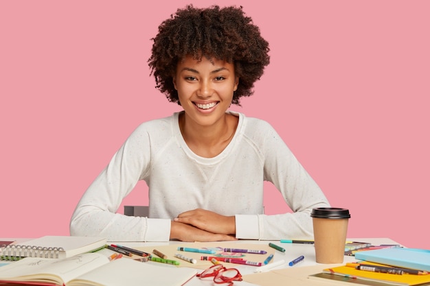 Isolierte Aufnahme von urkomischen dunkelhäutigen Frau hat Afro-Haarschnitt, gekleidet in lässigen weißen Pullover, sitzt am Schreibtisch mit kreativem Chaos, trinkt Kaffee zum Mitnehmen, verwendet Buntstifte, hat ein zahniges Lächeln im Gesicht