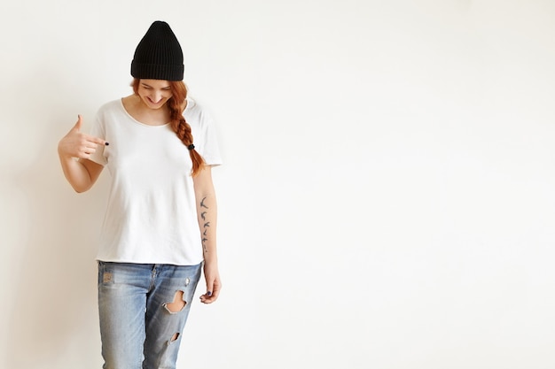 Kostenloses Foto isolierte studioaufnahme einer jungen frau mit zopf, die nach unten schaut, während sie auf ihr leeres weißes t-shirt zeigt
