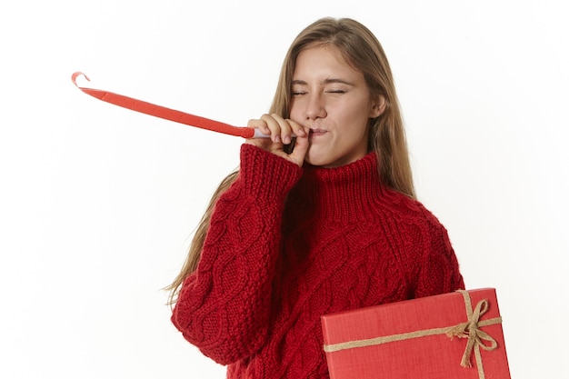Isolierter Schuss des emotionalen niedlichen weiblichen Teenagers, der stilvollen gestrickten Pullover aufwirft, der eine schicke rote Schachtel hält und Pfeife auf Geburtstagsfeier bläst, um ihrem Freund Geschenk zu geben