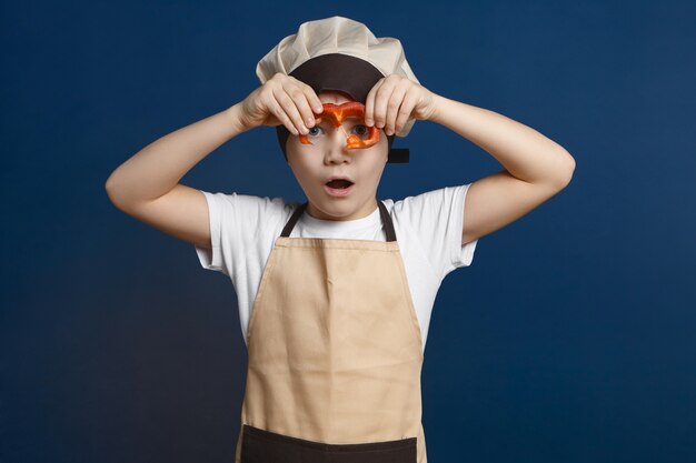 Isolierter Schuss des hübschen niedlichen kleinen Jungen in der Kochuniform, die Scheibe Paprika an seinem Gesicht hält