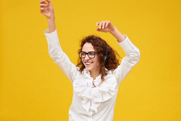 Isoliertes Bild der positiven emotionalen jungen Frau mit dem gelockten dunklen Haar, das an der gelben Wand mit den Händen oben in der Luft aufwirft, tanzt, Musik auf Kopfhörern hört, aufgeregt lächelt und Brille trägt