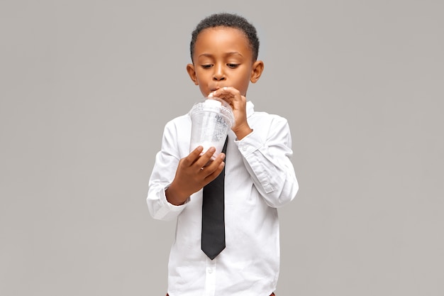 Isoliertes Bild des hübschen afrikanischen Schuljungen, der an der leeren Wand mit dem Plastikglas nippt, das Milchshake während der Mittagspause an der Schule nippt, erfreuten Blick. Lebensmittel-, Getränke- und Lifestyle-Konzept