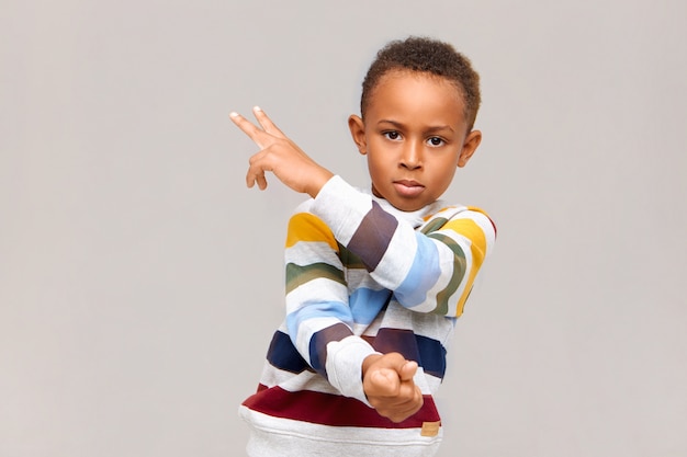 Isoliertes Bild des selbstbewussten gutaussehenden afroamerikanischen Jungen, der gestreiften Pullover trägt, der auf leere Wand gestikuliert, zwei Zeichen mit Fingern zeigend zeigt, rechts mit ernstem Blick starrend