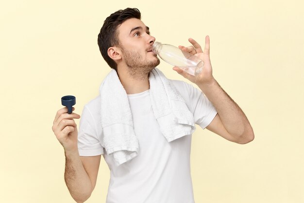 Isoliertes Bild des selbstbewussten gutaussehenden jungen Mannes mit Handtuch um den Hals, der Plastikflasche hält, sich nach körperlicher Übung im Fitnessstudio erfrischend, Wasser gierig trinkend, weißes T-Shirt tragend