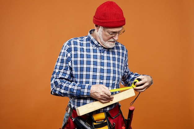 Isoliertes Bild eines ernsthaften bärtigen älteren männlichen Konstrukteurs in Brille und Hut mit fokussiertem Gesichtsausdruck, der Messungen der Holzplanke unter Verwendung des Maßbandes nimmt. Handarbeit und Arbeit